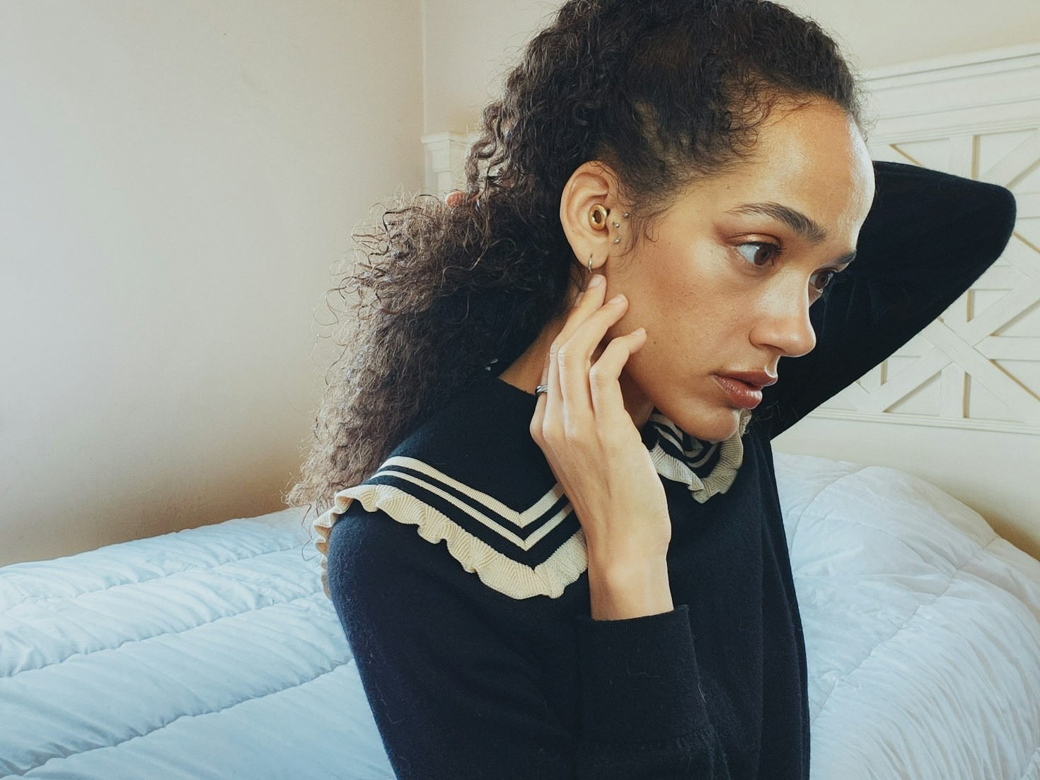 A person with curly hair adjusts an earbud while sitting on a bed with a blue comforter. They are wearing a black sweater with a distinctive collar featuring a white and black striped pattern and ruffled edges.