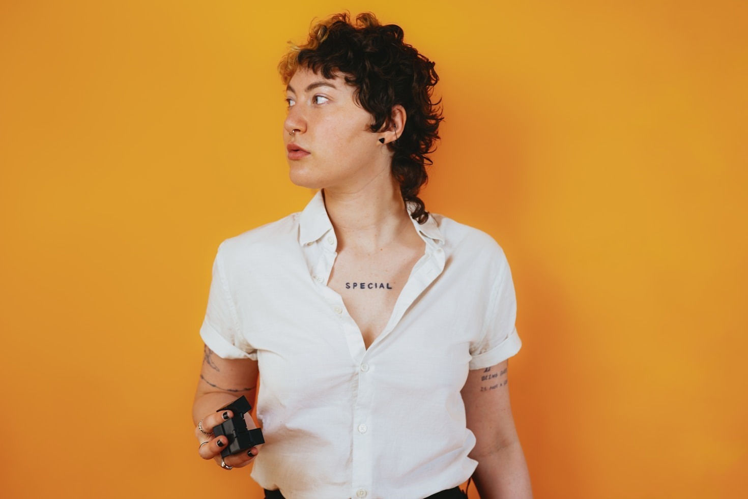 A person with short, curly hair stands against an orange background, wearing a white button-up shirt. The individual holds a small, black object in one hand and has tattoos, including one on the chest reading SPECIAL.