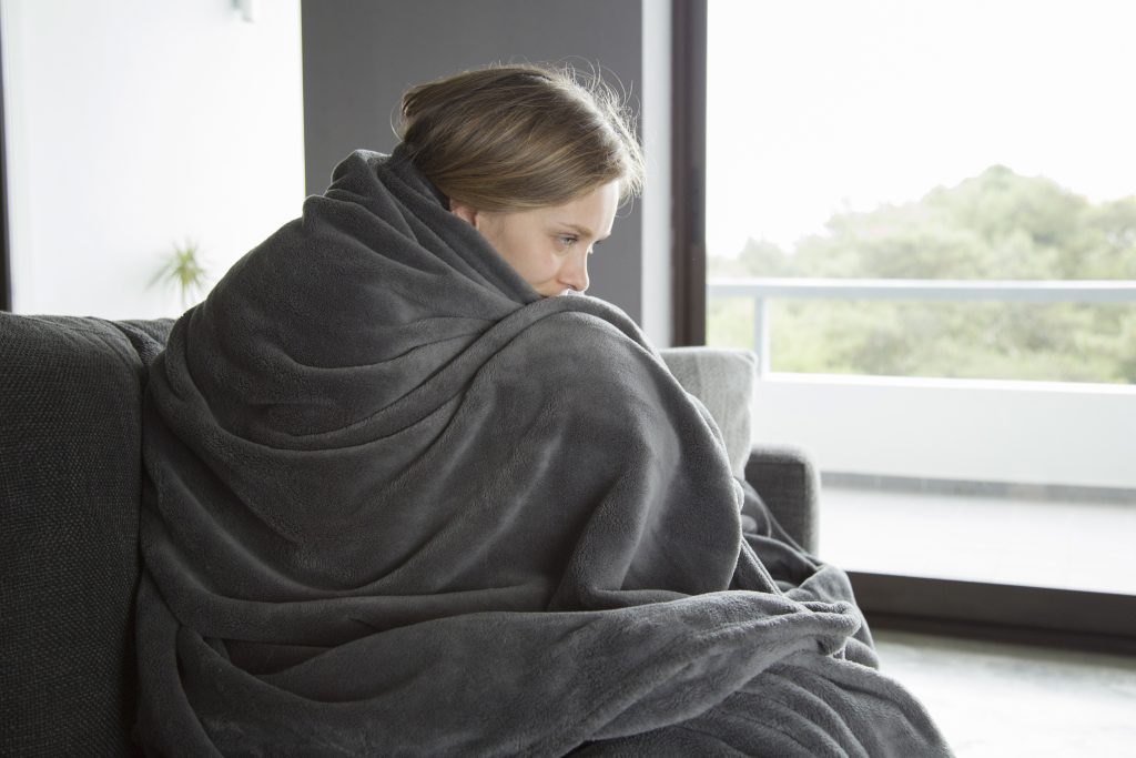 Young fair-haired Caucasian woman in purple sweater covered with grey blanket sitting on grey sofa in living room,
