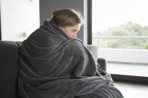 Young fair-haired Caucasian woman in purple sweater covered with grey blanket sitting on grey sofa in living room,