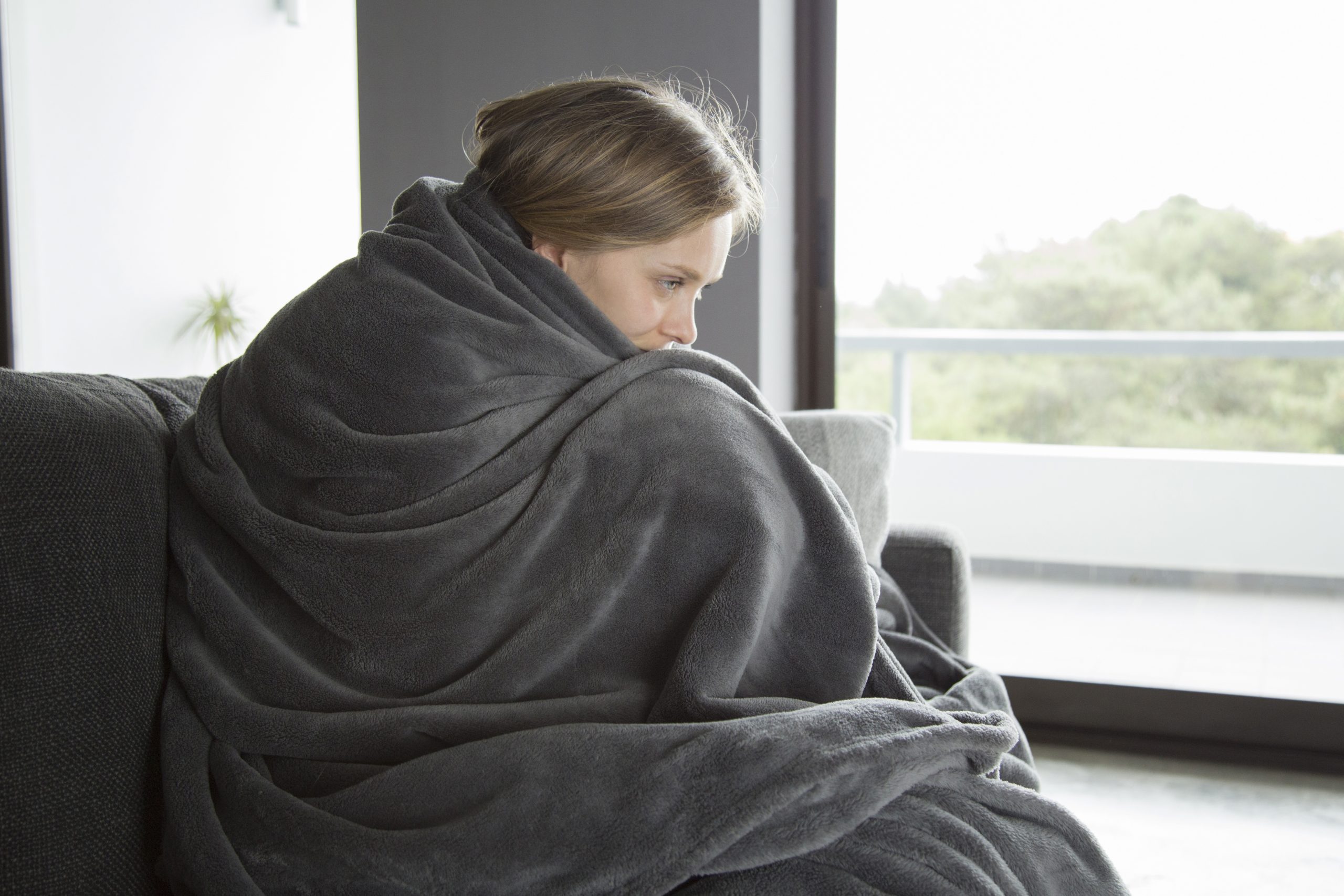Young fair-haired Caucasian woman in purple sweater covered with grey blanket sitting on grey sofa in living room,
